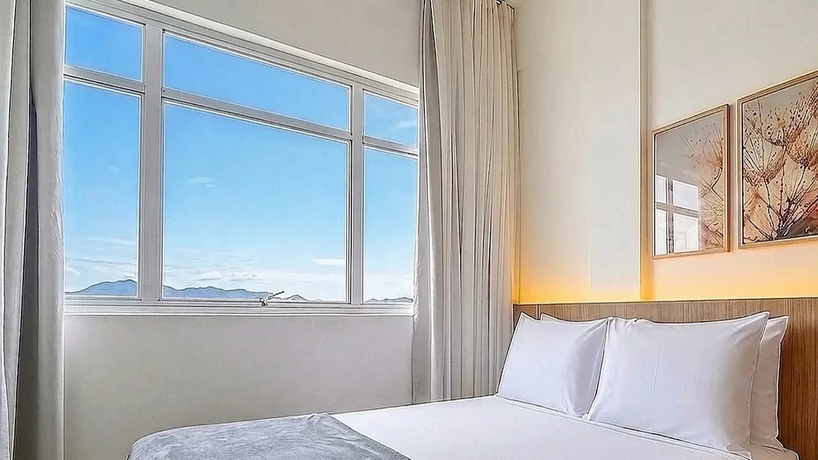 Cama de casal arrumada ao lado de uma janela ampla com vista para o céu